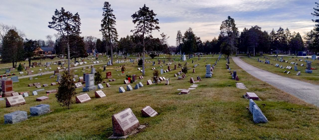 A large cemetery with numerous gravestones spread across a grassy area. A paved path winds through the site, and mature trees are scattered throughout. The sky is cloudy, casting a serene atmosphere over the peaceful landscape.