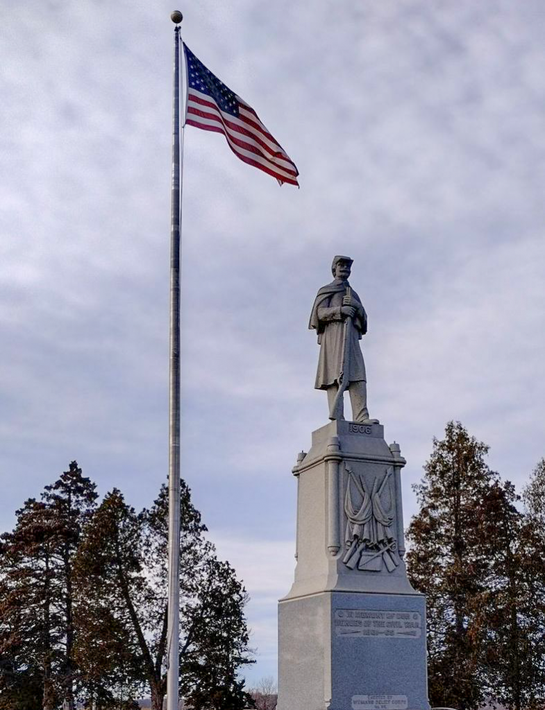 A tall monument depicting a soldier stands on a pedestal adorned with carved decorations, under a partly cloudy sky. Next to the monument is a flagpole with the American flag unfurled. Trees with sparse foliage are visible in the background.