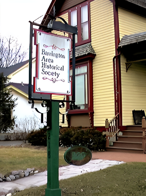 A two-story yellow and red building with a porch. In front of the building, there is a tall, green metal signpost with a white sign that reads "Barrington Area Historical Society." The surrounding area includes grass, bushes, and a paved pathway.