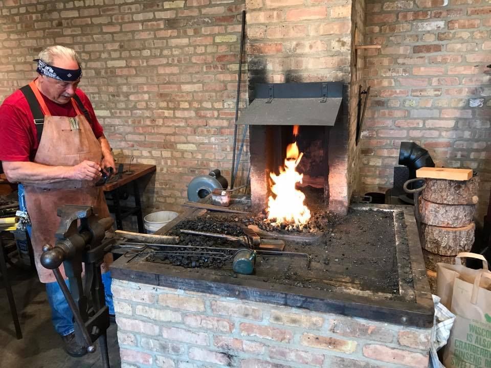 A blacksmith wearing a red shirt, tan apron, and black headband works in a rustic workshop. He stands near a blazing forge with various tools and materials scattered around. The workshop features brick walls and a large chimney for the forge.