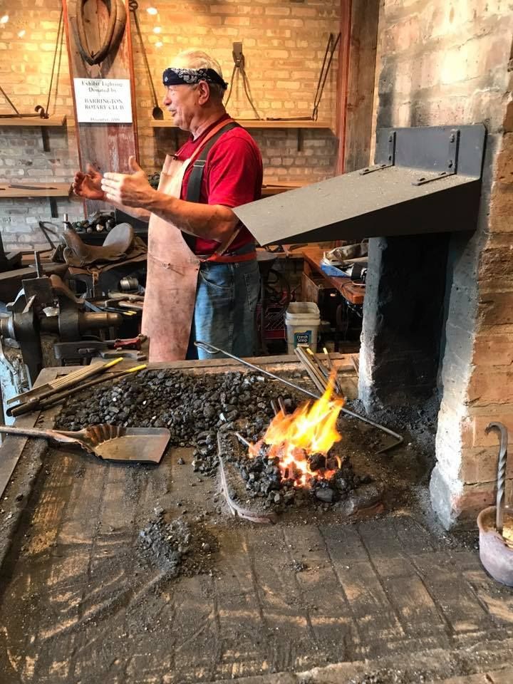 A blacksmith stands in a workshop, wearing a red T-shirt, jeans, a leather apron, and a patterned bandana. There is a burning forge with coal in the foreground and various blacksmithing tools scattered around. The workshop has brick walls and industrial decor.