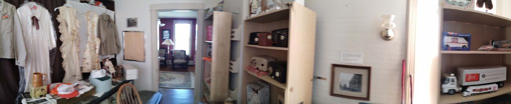 Wide-angle image showing rooms with clothing hanging against the left-side wall. On the opposite wall are three shelving units with toy vehicles from the Jewel Tea Company