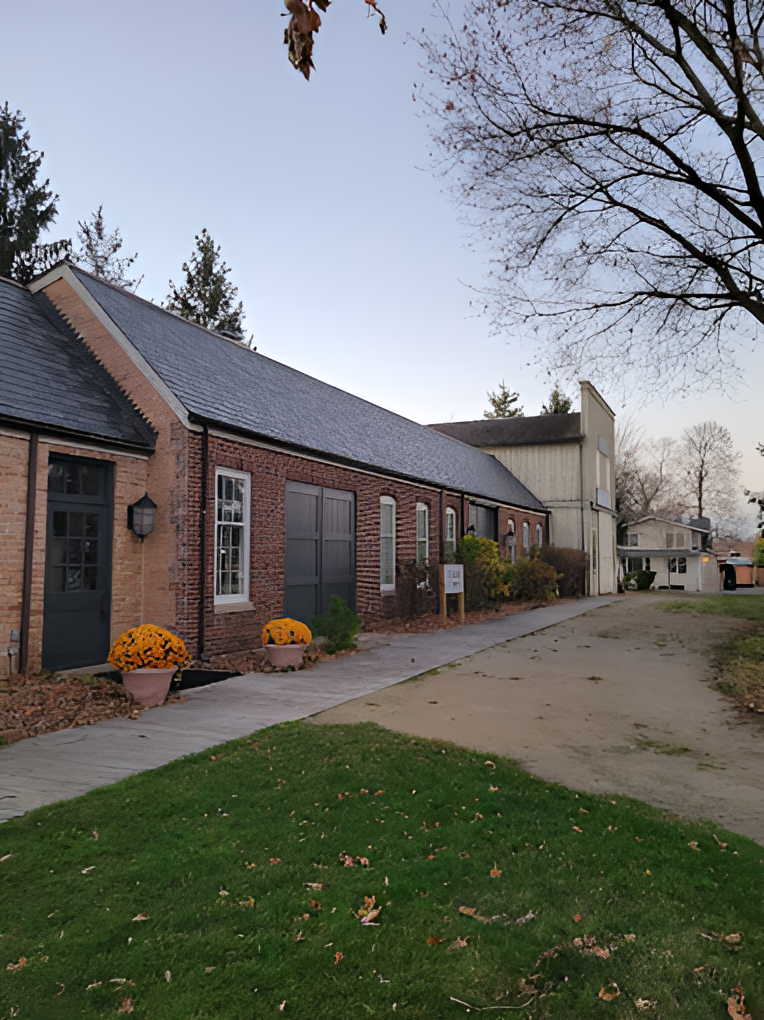 Brick building with grey roof surrounded by trees and a green lawn. Orange flowers and walkway in front. Dusk sky visible.