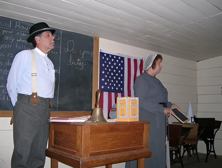 A man and a woman dressed in historical attire stand in a vintage classroom. The man wears a hat and suspenders, and the woman wears a bonnet and long dress. An American flag and chalkboard are visible in the background. A wooden desk with a bell is in the foreground.