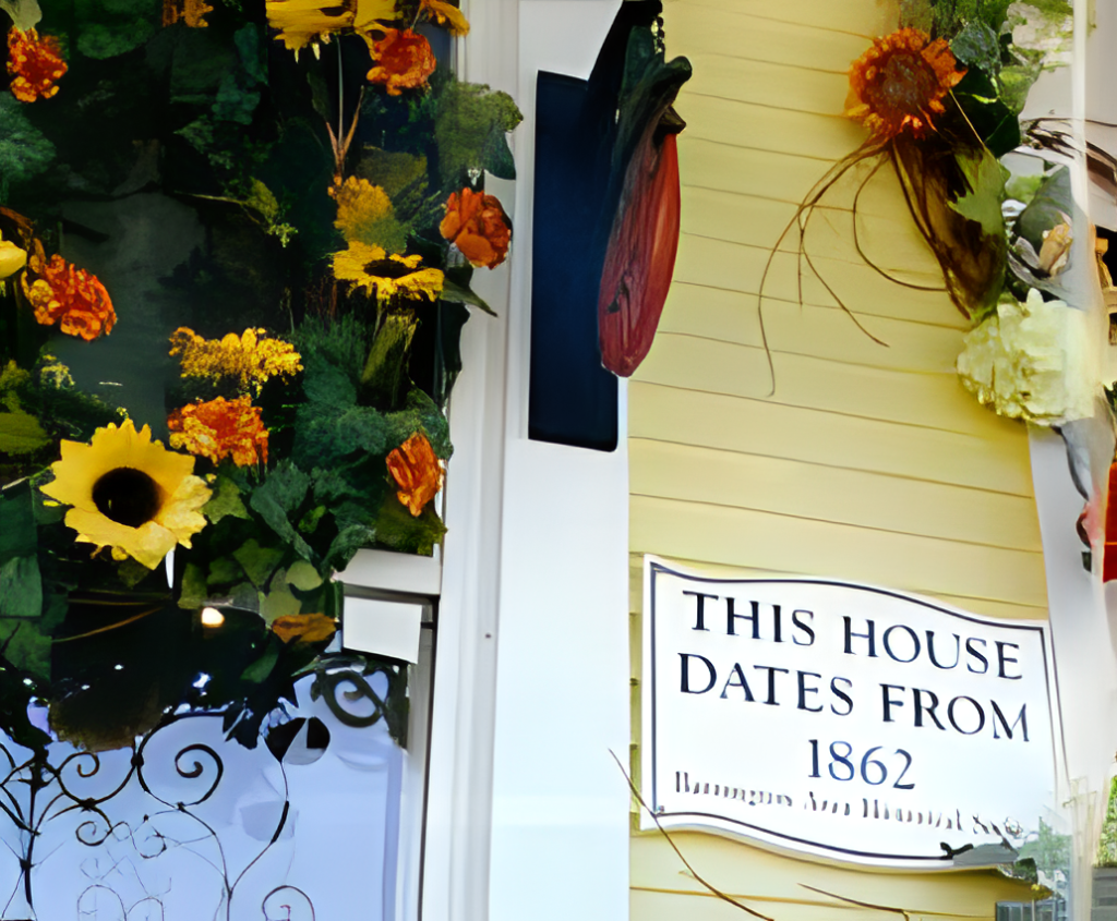 A historic house sign reads "THIS HOUSE DATES FROM 1862" against a yellow wall. The surrounding decor features vibrant, colorful flowers, including sunflowers and various other blooms, adding a cheerful atmosphere to the scene.