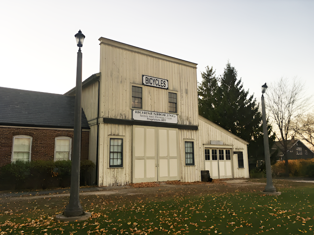 A historical building featuring a sign that reads "Bicycles" on the top and "Horse Shoeing" on the bottom. The structure is flanked by two vintage-style lampposts and surrounded by scattered autumn leaves. Trees and additional buildings are visible in the background.