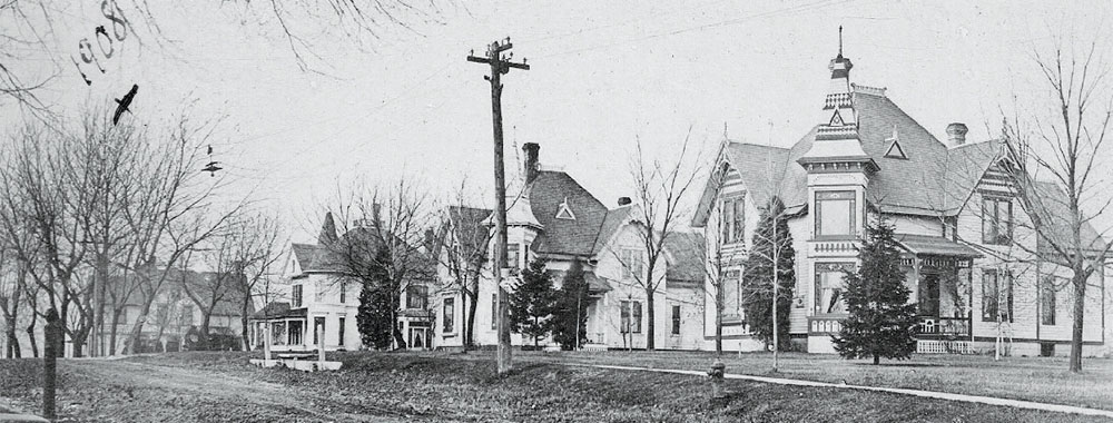 An old photograph of 3 victorian houses. Electric poles and trees are visible. 1908 has been written on top of the photo.