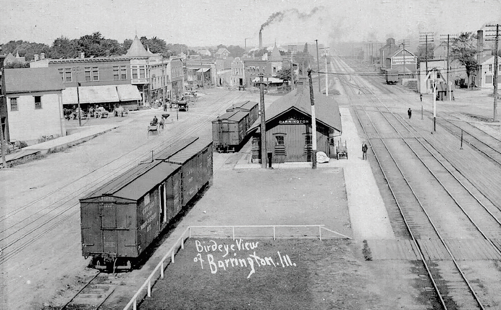 Black and white birdseye photograph of a building with train tracks on both sides. The track on the left has several train cars. Smoke is billowing from chimneys. People and buildings are in the background.