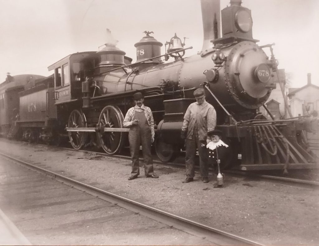 A black and white photo of a steam locomotive labeled "678" at a train station. Three individuals stand in front of it: two adults in overalls and a child wearing a suit and hat. The adults appear to be railway workers.