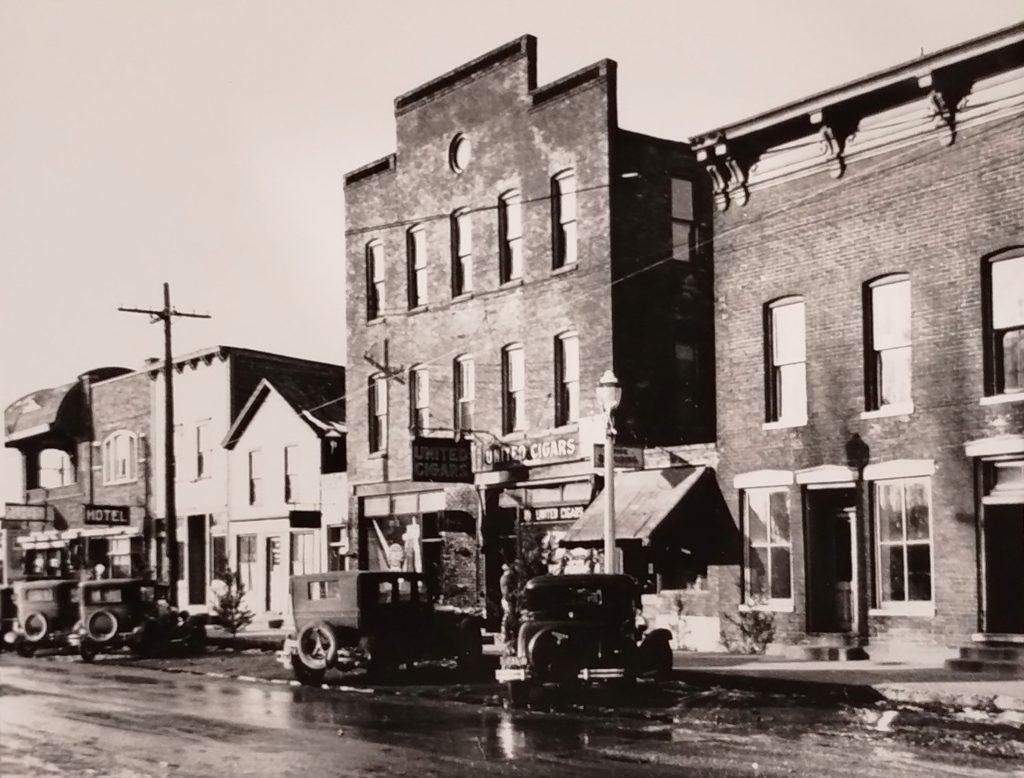 Black and white photo of a historic street scene with several brick buildings, including a hotel and a cigar store. Vintage cars are parked along the wet road. The buildings have arched windows and decorative cornices, indicative of early 20th-century architecture.