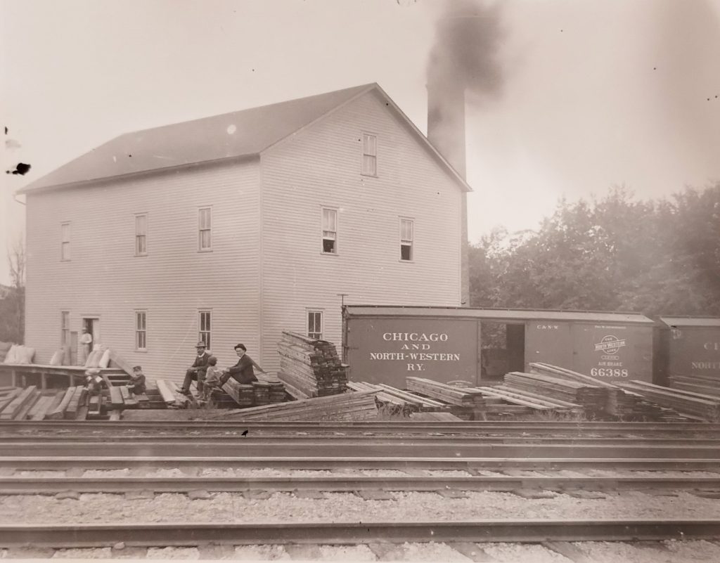 Black and white image of a building near railroad tracks. Several men sit on stacks of wooden planks beside two train boxcars labeled "Chicago and North-Western Ry." Smoke rises from a chimney in the background.