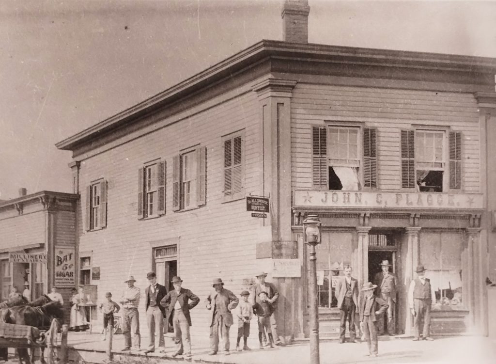 A sepia-toned historical photo of a two-story building labeled "John C. Plagge" with men and boys gathered outside. The building features large windows with shutters and a notable sign for "Bat 5¢ Cigar." Some signs are in the windows and on the walls.