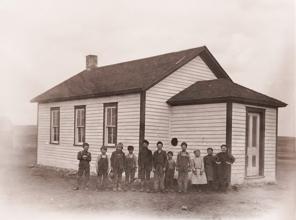 A sepia-toned photograph shows a group of children standing in front of a small, single-story, wooden schoolhouse with a gabled roof. The children are dressed in early 20th-century clothing and pose in a line facing the camera.