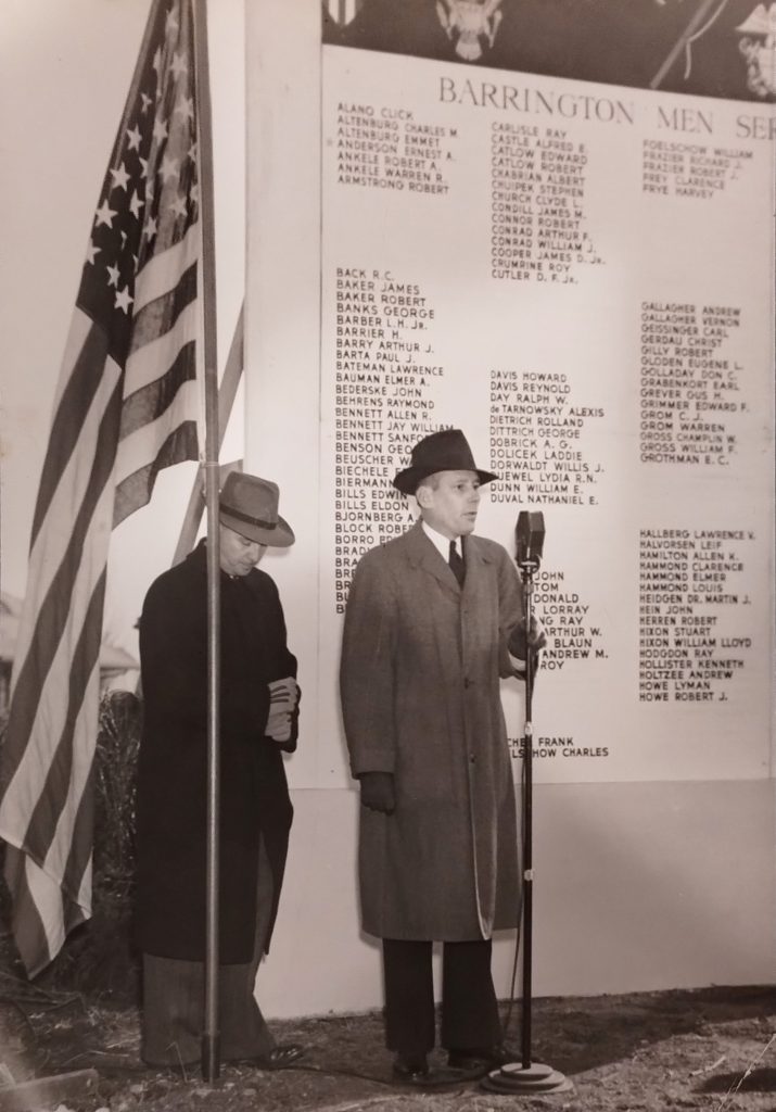 A man is speaking at a microphone with a man standing behind him. On the left is a flag pole with an American flag, and on the right is a monument featuring a list of names.