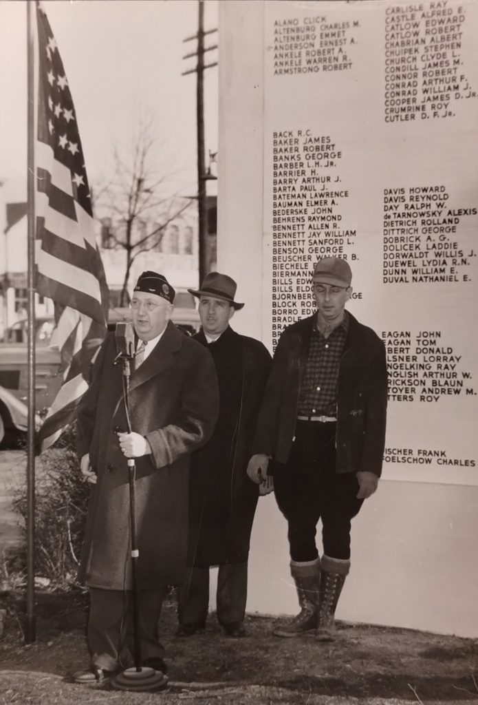 A man is speaking at a microphone and two men are standing behind him. On the left is a flag pole with an American flag. On the right is a monument featuring a list of names.